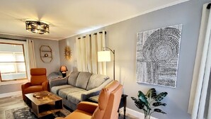 A modern living room with a grey sofa, orange armchairs, a coffee table, and a floor lamp. - The Gorgeous George Beach Cottage (East Haven)