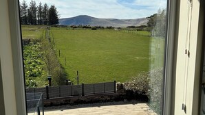 Une vue depuis une fenêtre donnant sur un champ verdoyant, avec un mur de pierres et une terrasse en bois.