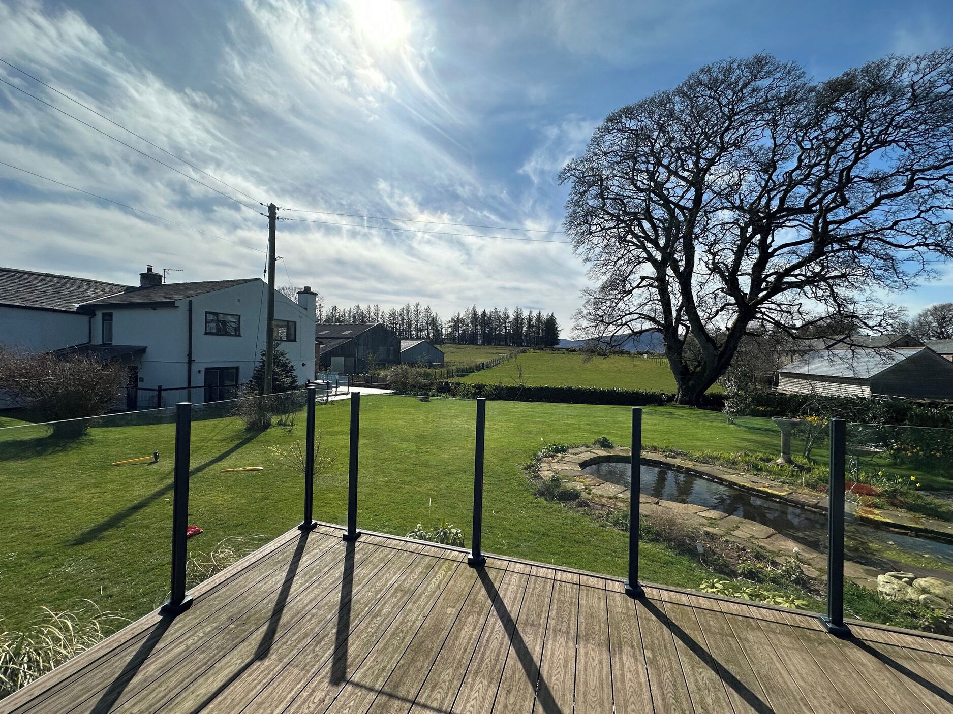 A wooden deck with a view of a grassy yard, a pond, and a large tree.