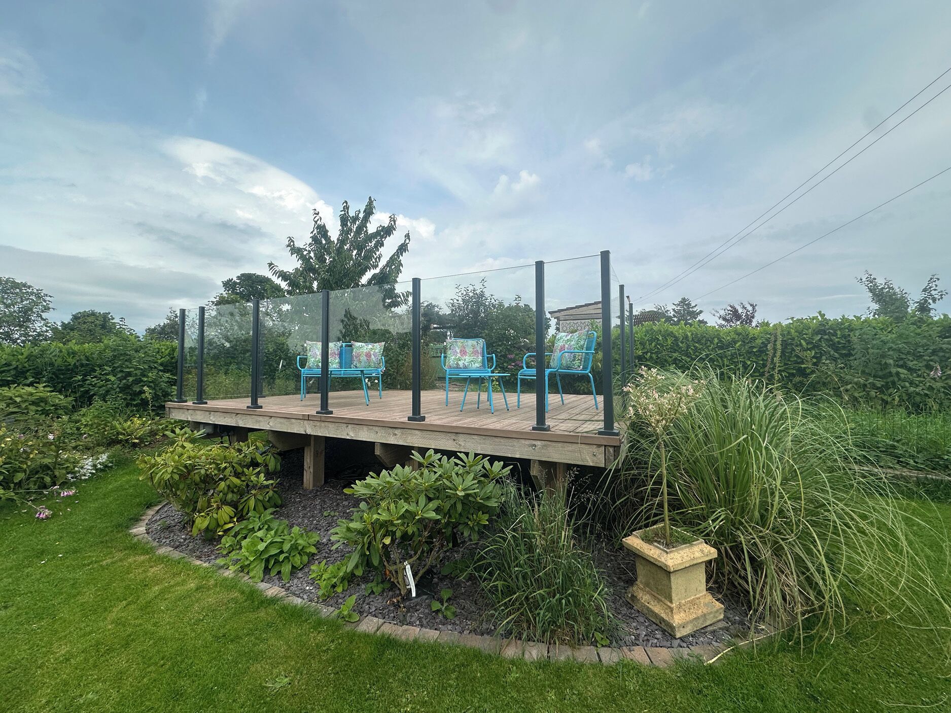 Um deck de madeira com cadeiras azuis, cercado por um jardim com diversas plantas e um vaso de pedra.