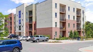 A multi-story residential building with balconies, parking spaces, and a clear sky. - 1st Floor Condo | 2 Queen Beds + TART Trail (Traverse City)