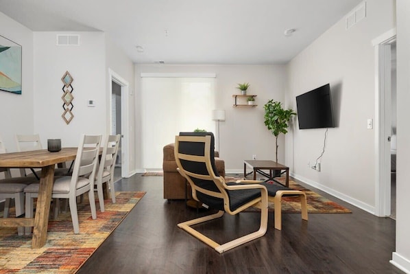 A modern living room with a dining area, a TV mounted on the wall, and a large window providing natural light. - Trailside Condo • 2BR/2BA • 1.5mi to DT (Traverse City)