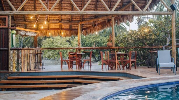 Un espace extérieur avec une piscine, des sièges en bois et une structure dotée d’un toit en chaume.