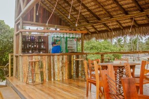 Uma área de bar ao ar livre com estrutura de madeira, balcão com garrafas e assentos.