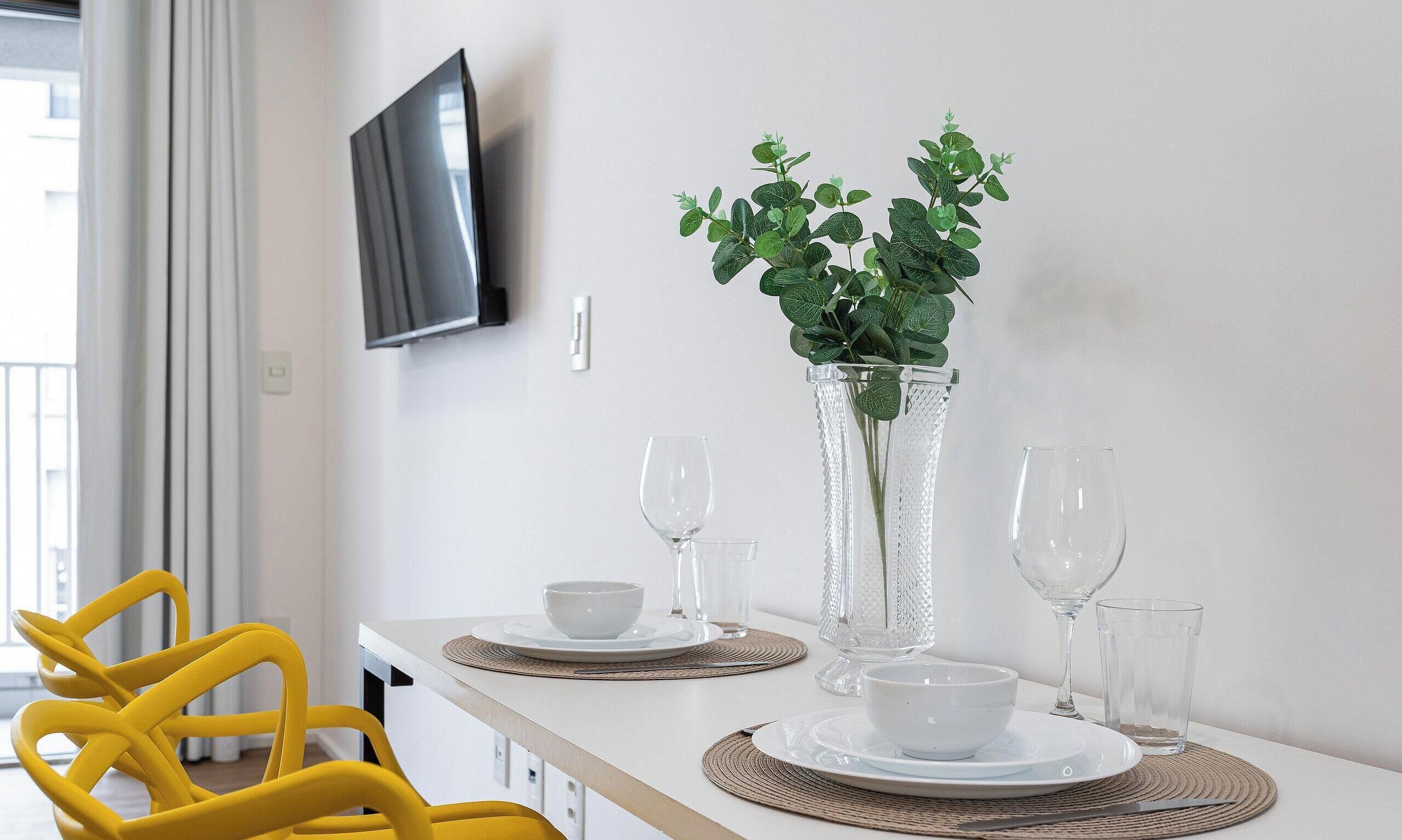 A dining area with a white table set for two, a yellow chair, and a potted plant on the table.