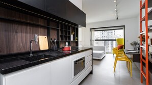 A modern kitchen with a black countertop, stainless steel appliances, and a red pot. There is a dining area with a yellow chair and a red bookshelf. - Aurora Paulistana by Viva - Prédio completo a passos do Metrô República (São Paulo)