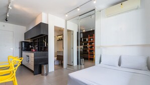 A modern bedroom with a large bed, a small kitchen area, and a sliding glass door leading to a closet. - Aurora Paulistana by Viva - Prédio completo a passos do Metrô República (São Paulo)