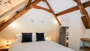 A bedroom with a high ceiling and exposed wooden beams, a bed with white and black bedding, and a nightstand with a lamp. - Eventide (Plymouth)
