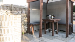 A wooden pergola with a table and benches, two wine glasses, and bottles on the table. - Eventide (Plymouth)