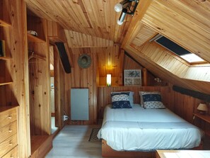 Une chambre confortable aménagée dans une cabane en bois, avec un plafond en pente, un lit agrémenté d’oreillers sur le thème de la marine, un petit bureau et une lucarne.