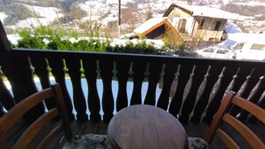 A balcony with a wooden chair and a small table, overlooking a snowy village with traditional houses and a mountain in the background.