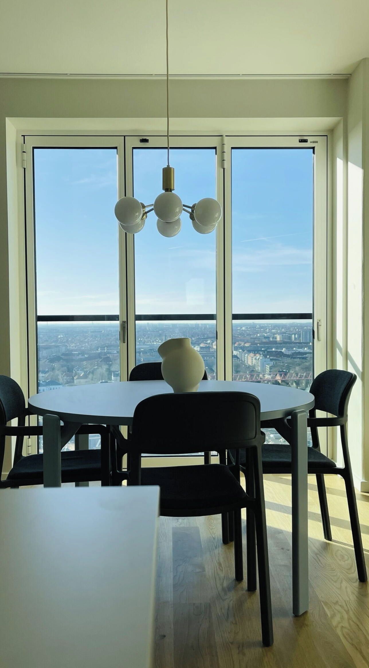A dining area with a table and chairs, a vase on the table, and a city view through large windows.