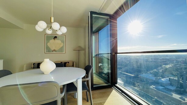 Ein moderner Essbereich mit einem runden Tisch, Stühlen und einer Vase. Ein gerahmtes Kunstwerk hängt an der Wand, und ein großes Fenster bietet Blick auf die Stadt.