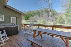 Relax on the deck with a hot tub, wooden table, and two comfortable rocking chairs overlooking the serene natural surroundings.