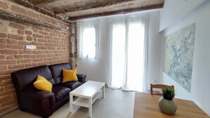 A living room with a brick wall, a brown leather sofa with yellow cushions, a white coffee table, and a wooden dining table with a plant. - Charming Loft in the Historic Center of Manresa. (Manresa)