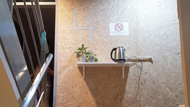 A staircase with a metal railing leading to a landing. On the landing, there is a wall-mounted shelf with a plant and a kettle. A 'No Smoking' sign is visible on the wall. - Foresttel Bkk - Hostel (Bangkok)