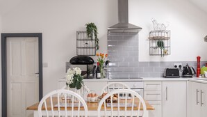 Een moderne keuken met witte kasten, een ingebouwde kookplaat en een roestvrijstalen afzuigkap. Er staan twee witte stoelen met houten zitvlakken, een kleine tafel en staat er een potplant op het aanrecht.
