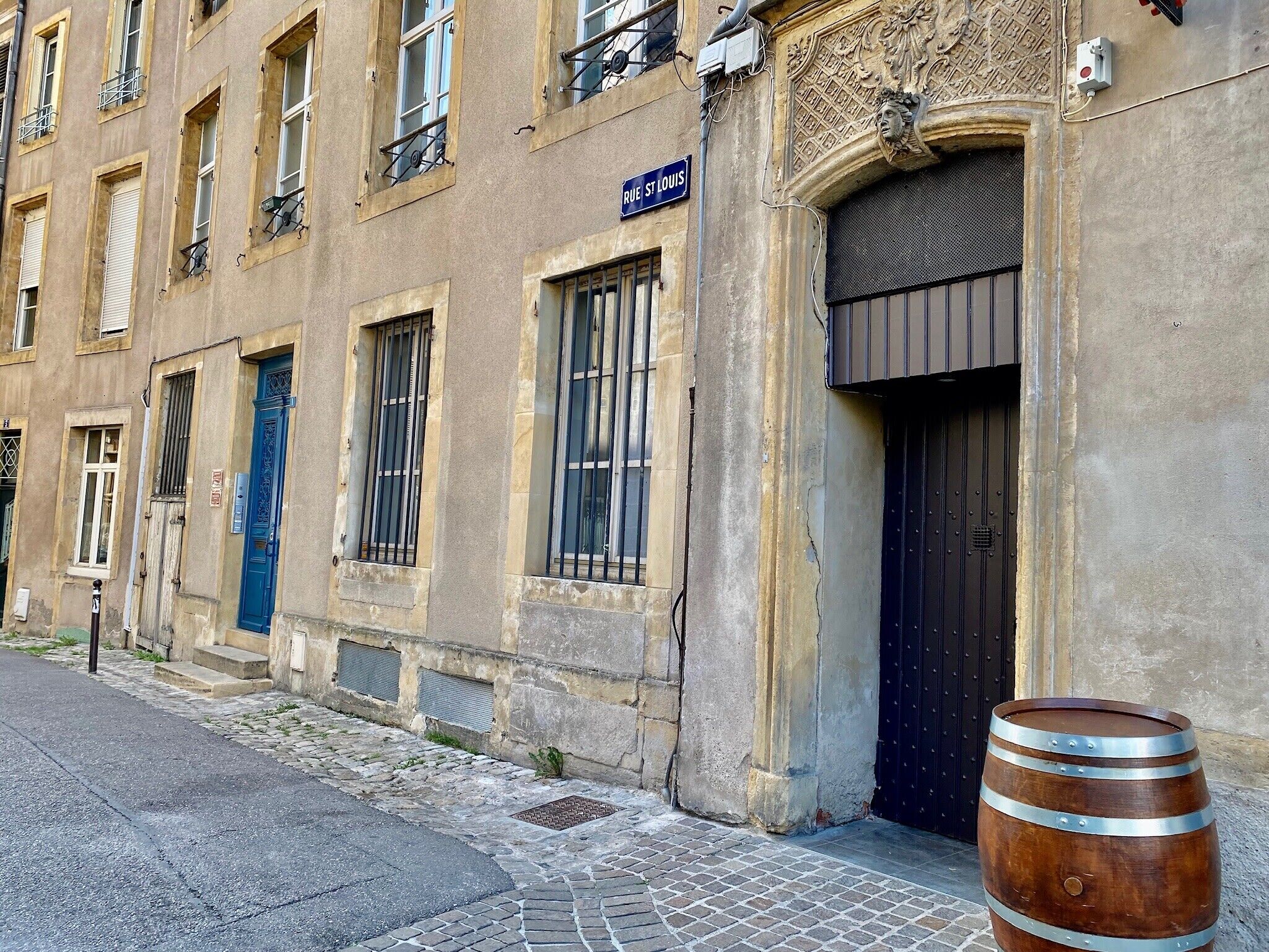 A building with a blue door, a sign reading "Rue St Louis," and a large barrel on the sidewalk.
