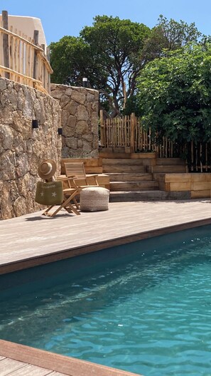 Un espace aménagé au bord de la piscine, avec un muret en pierre, des marches en bois et du mobilier d’extérieur comprenant une chaise et une petite table.