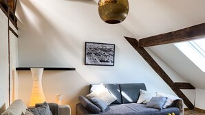 Un salon chaleureux de style loft, avec un plafond haut, des poutres apparentes, un canapé moderne, une table agrémentée d’un vase de fleurs et, au mur, un tableau représentant un paysage urbain.