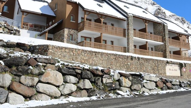 A multi-story hotel with stone walls and wooden balconies, set against a snowy mountain backdrop.