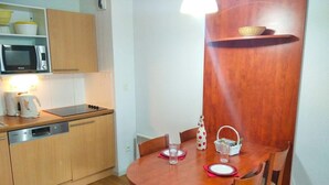 A compact kitchen with a microwave, dishwasher, and wooden cabinets. A dining table with chairs is positioned under a wooden beam, and a small shelf holds a basket.