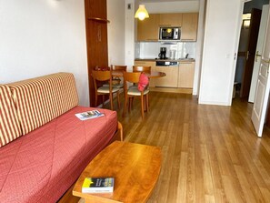 Um espaço compacto com sofá, uma pequena mesa de madeira, área de cozinha com micro-ondas e cafeteira, e mesa de jantar com cadeiras.