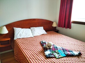 Uma cama com colcha xadrez, duas almofadas brancas, um urso de pelúcia, um cobertor e uma mesa de cabeceira com uma luminária.