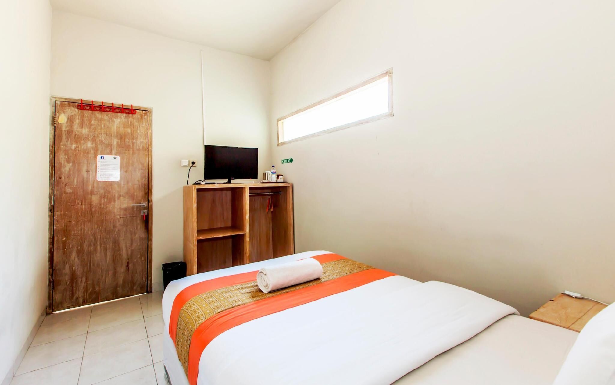 Una habitación pequeña y limpia con una cama, un armario de madera, un televisor sobre un mueble bajo y una ventana.