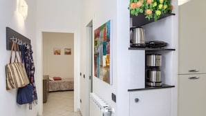 Un pasillo con un radiador, una cama con colcha estampada, un cuadro en la pared y una planta colgante con flores.
