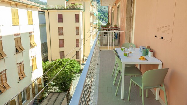 Un balcón con mesa y sillas, con vista a una calle con edificios y áreas verdes.