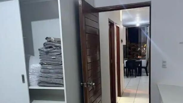 Um corredor estreito com uma cama à esquerda, um guarda-roupa com roupas dobradas e uma porta que dá para outro quarto.