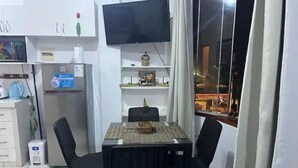 Espaço compacto com mesa de jantar, duas cadeiras, TV na parede, geladeira e janela com vista da cidade à noite.