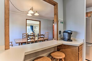 Uma cozinha moderna com bancada branca, cadeiras de madeira, cafeteira e geladeira.