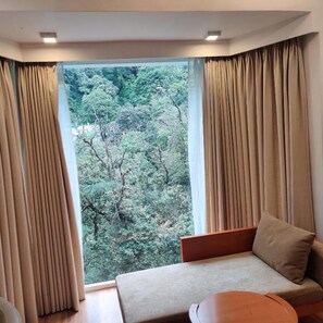 A room with a large window, beige curtains, a sofa, and a wooden table.