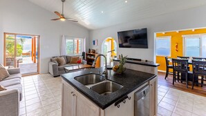 A modern kitchen with a central island, a dining area with a table and chairs, a living room with a sofa, and a TV mounted on the wall. - Blue Moon Villa (Tortola)