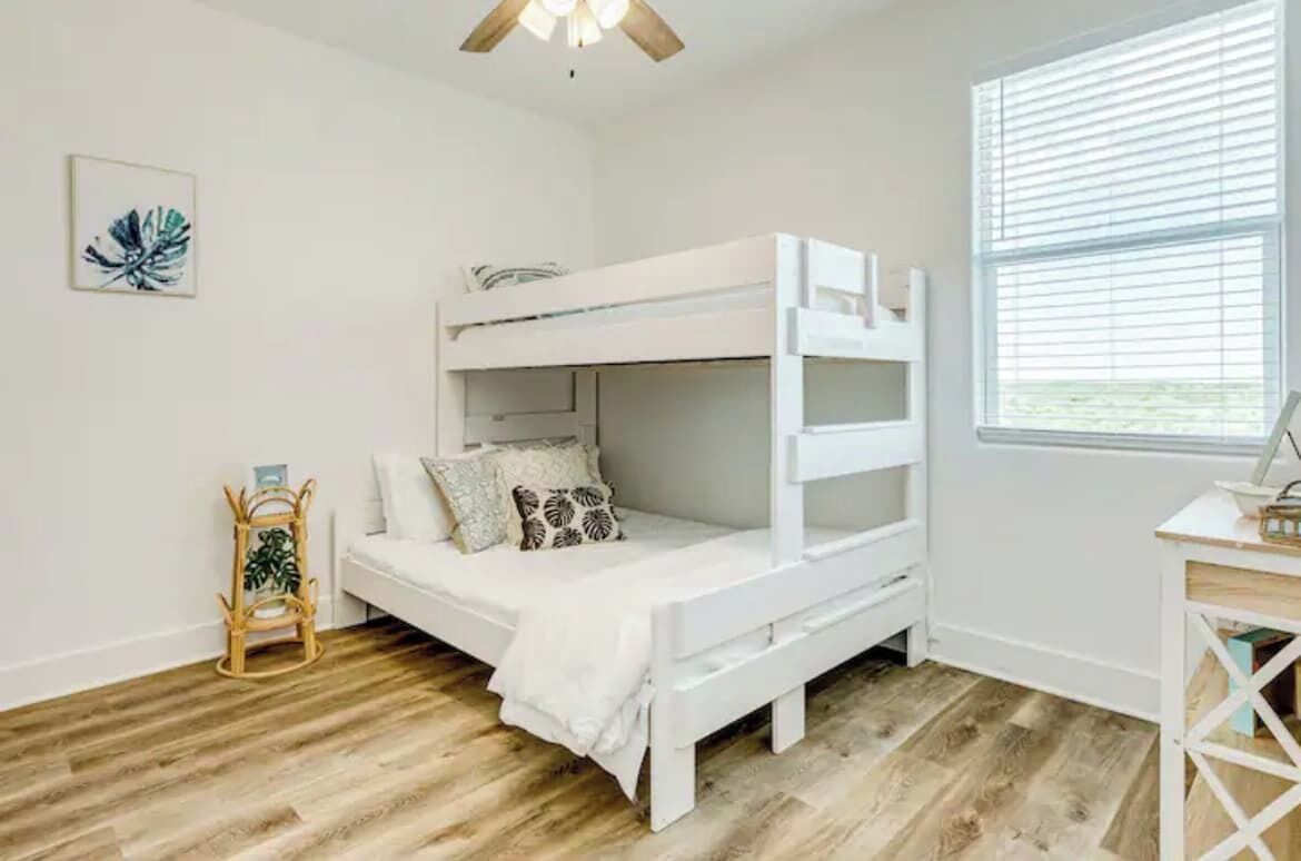 Een stapelbed in een kamer met houten vloeren, een bureau met stoel en een raam met zonwering.