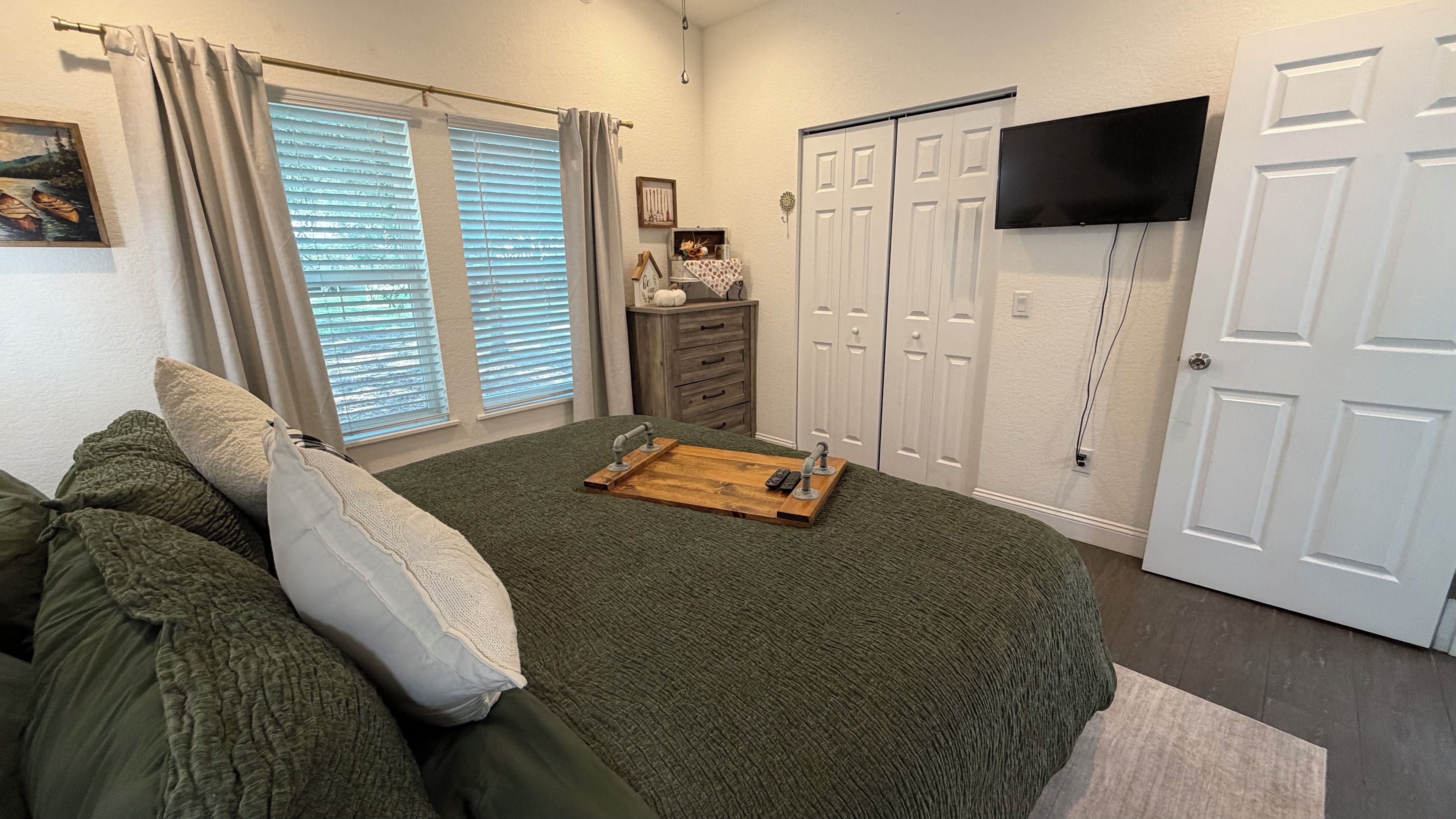 A bedroom with a bed, a dresser, a TV mounted on the wall, and a window with blinds.