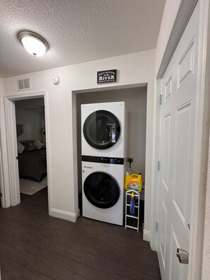 A laundry room with a stacked washer and dryer, a sign above the dryer, and a small shelf with cleaning supplies.