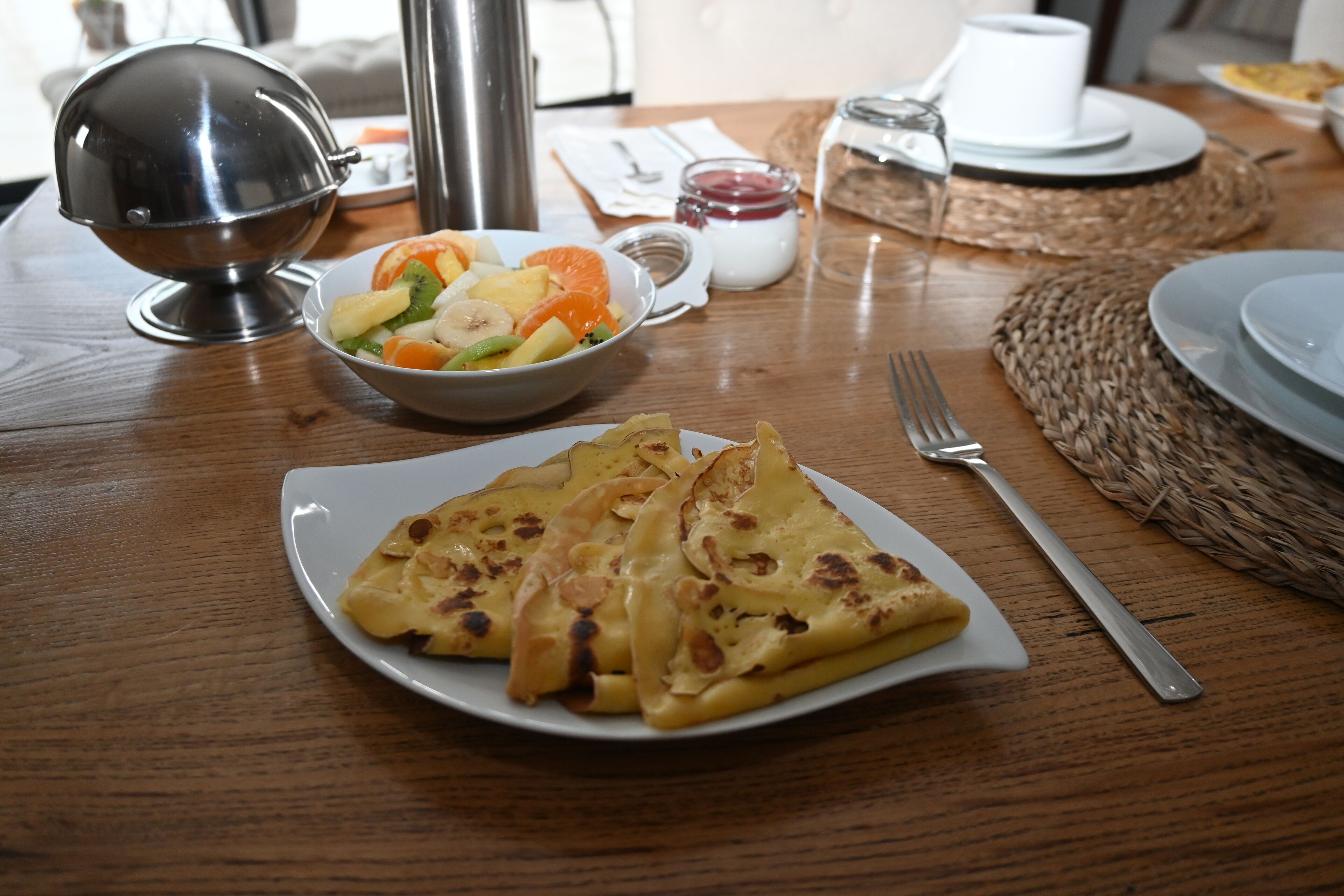 Een houten tafel met een stapel pannenkoeken, een schaal fruitsalade en een thermoskan.