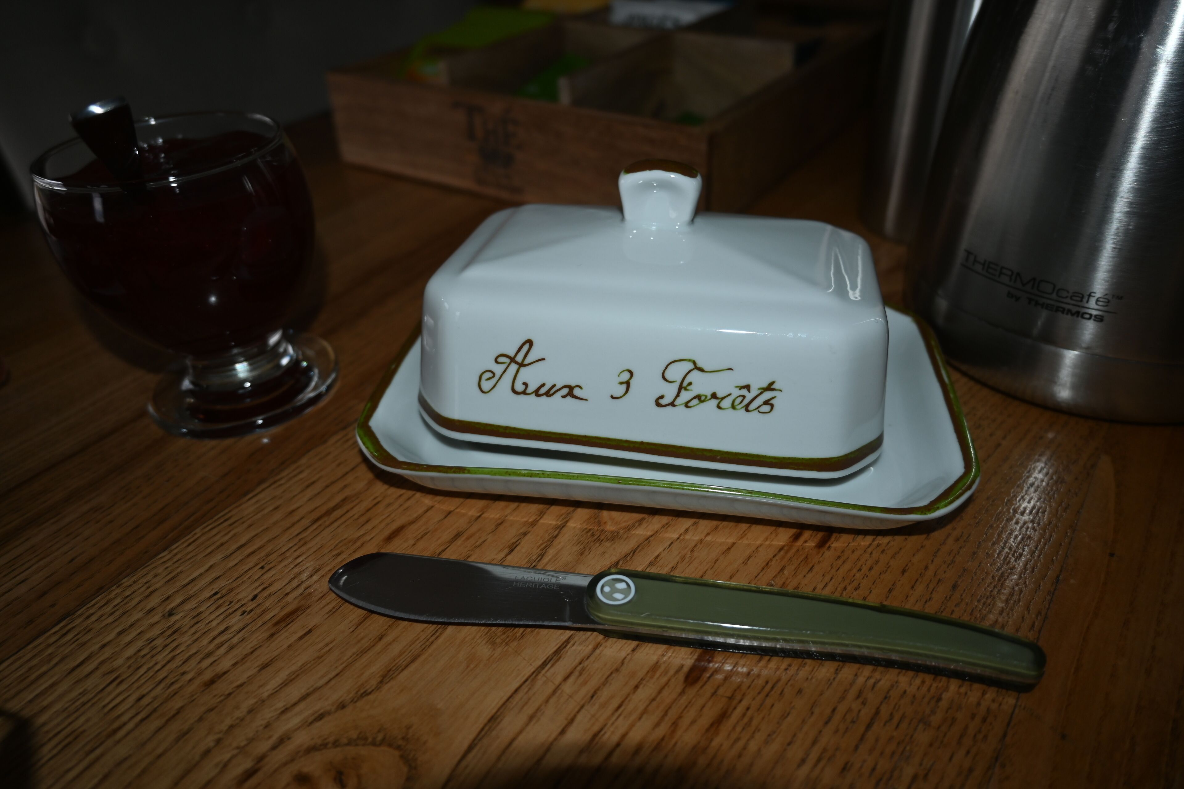 A white dessert with 'Aux 3 Fruits' on a plate, a knife, a glass, and a thermos on a wooden table.