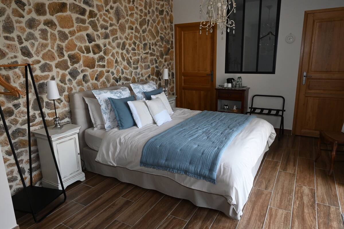 Une chambre avec un mur en pierre, un lit recouvert d’une couverture bleue, une table de chevet avec une lampe, une porte en bois et un lustre.