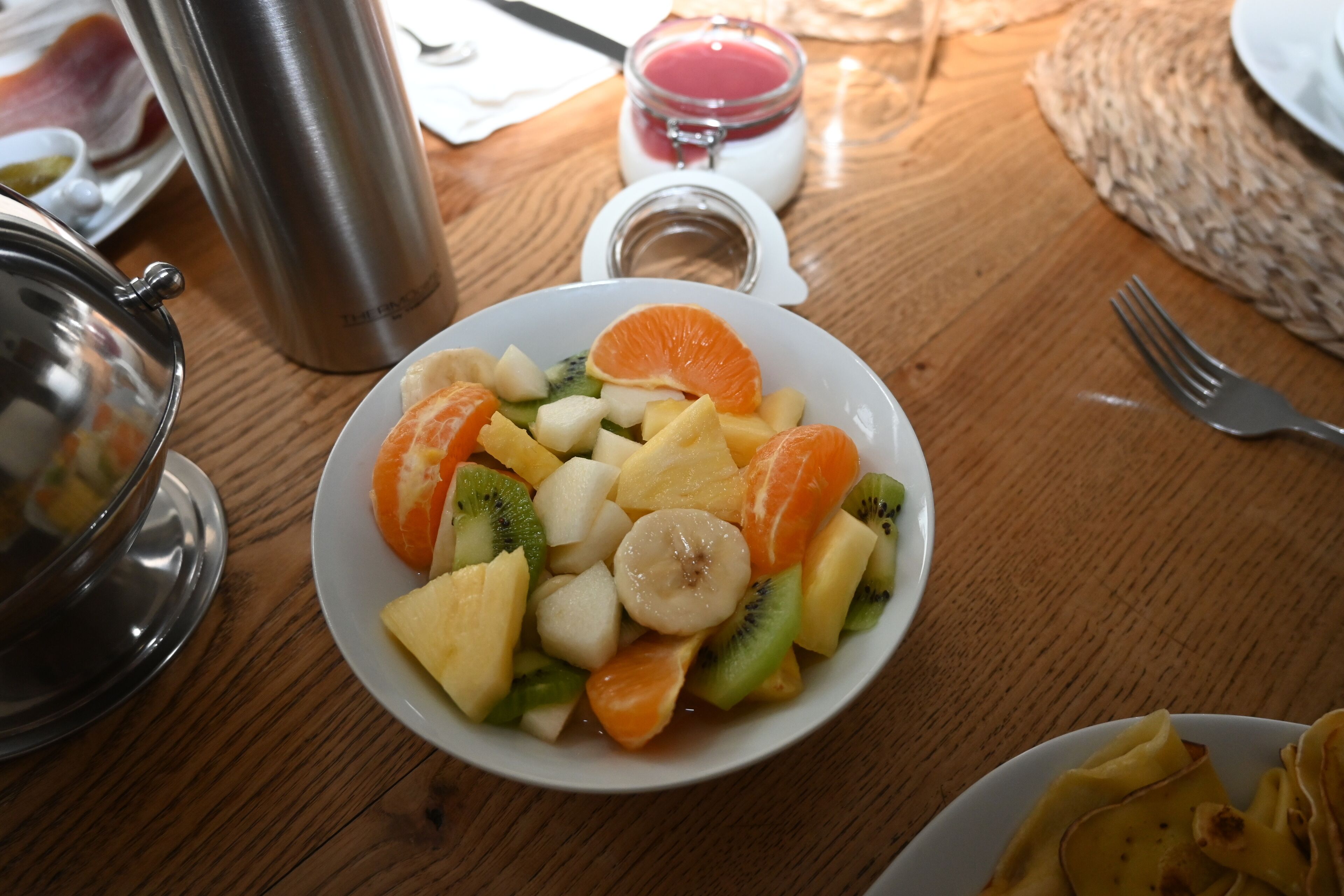 Una mesa de madera con un plato blanco de ensalada de frutas, un termo de acero inoxidable y un tarro de vidrio con un líquido rojo.