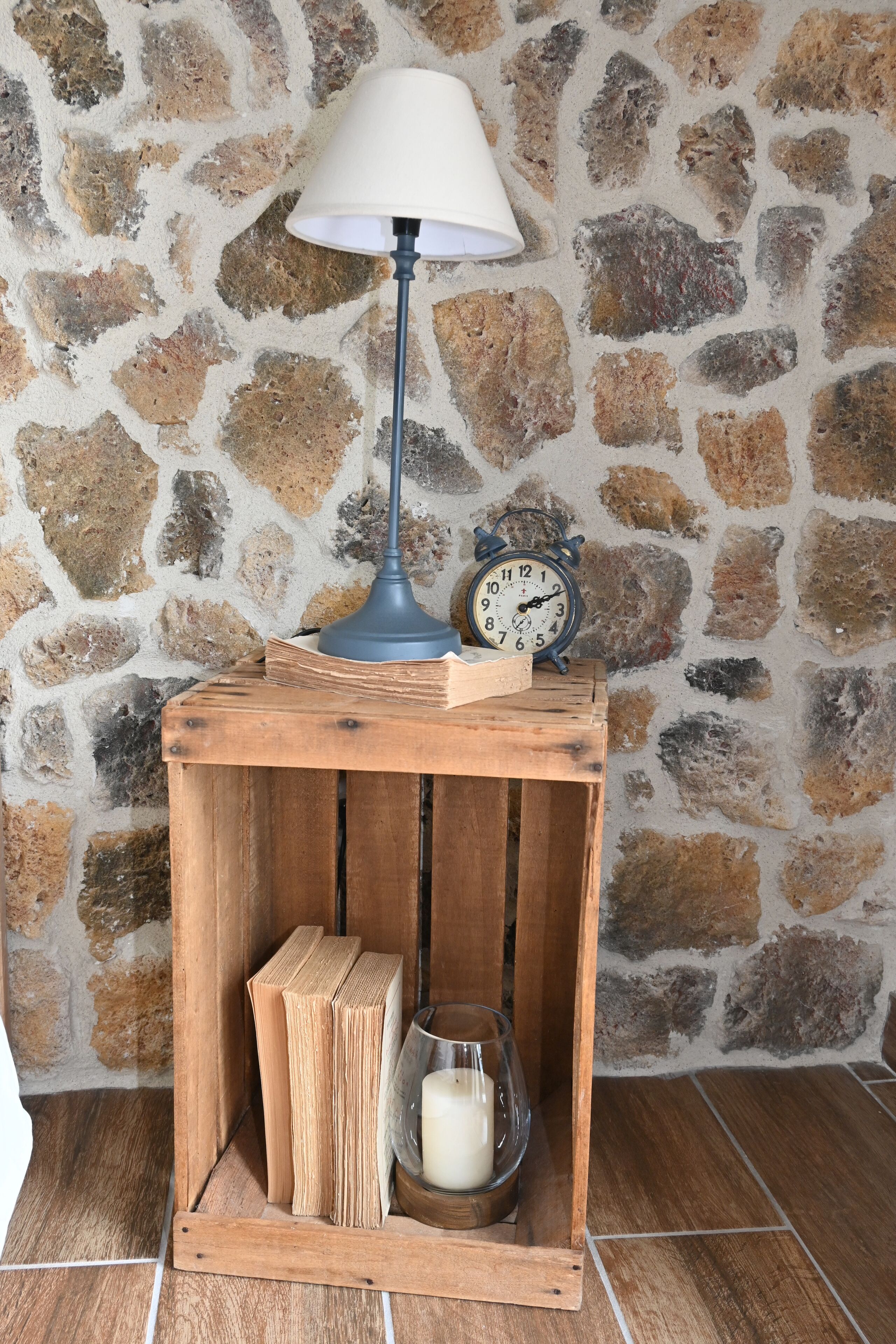 Een rustieke houten nachtkastje met een lamp, boeken en een kaars voor een stenen muur.
