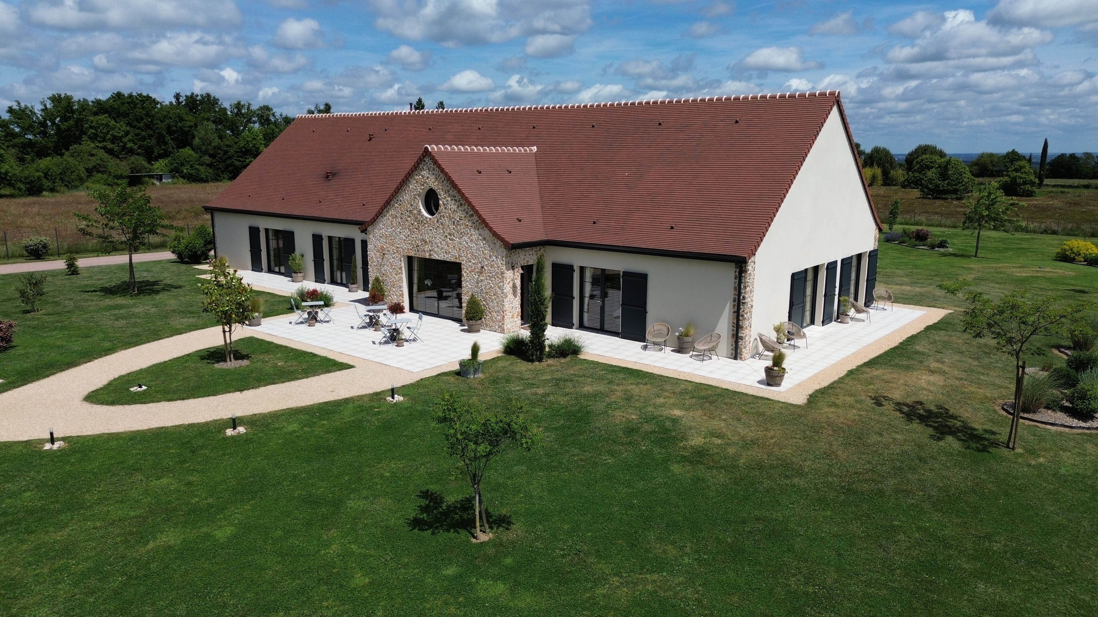 A modern two-story house with a stone facade, a red-tiled roof, and a landscaped garden with outdoor seating.