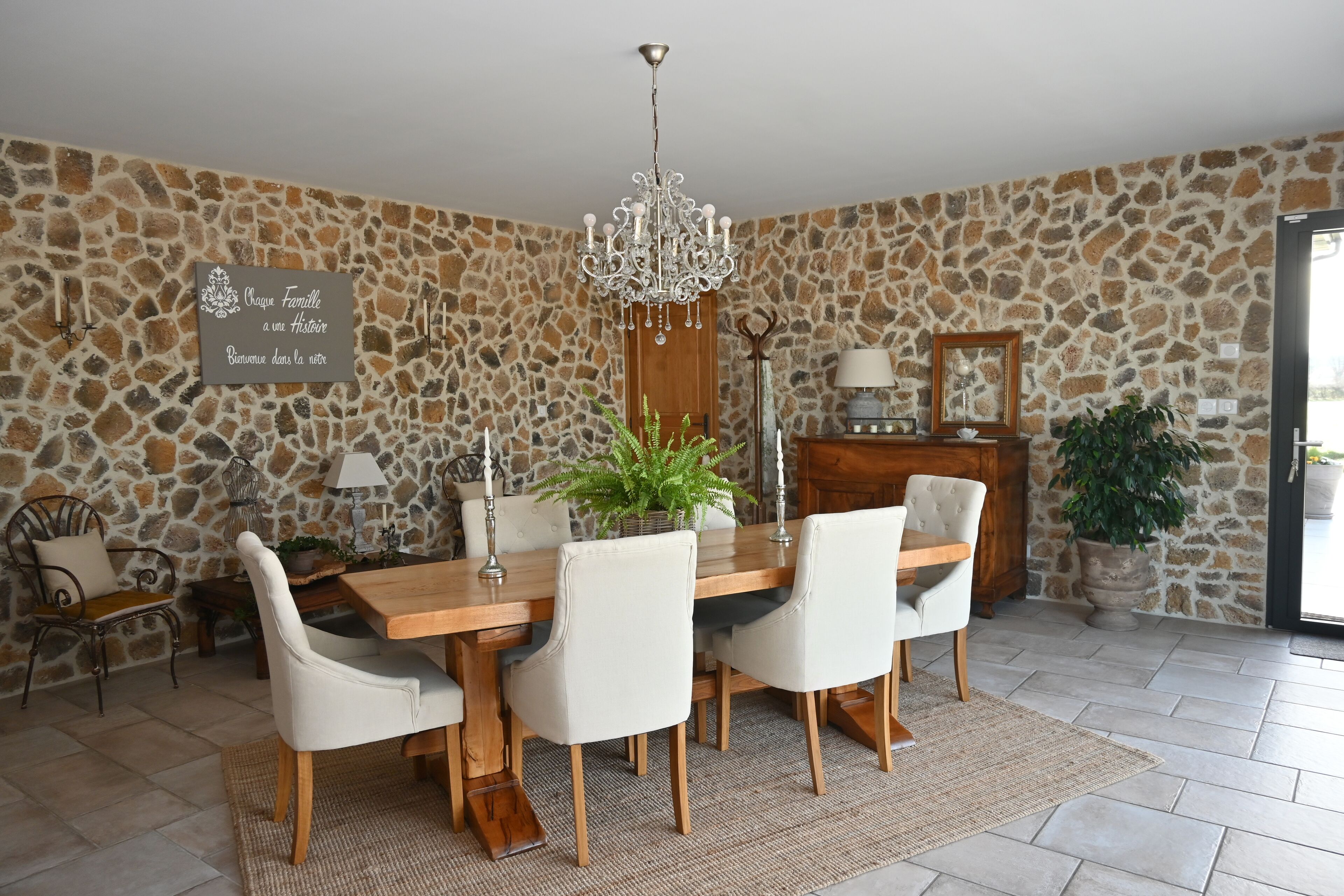 Un espace repas avec un mur en pierre, un lustre, une table en bois et des chaises blanches.