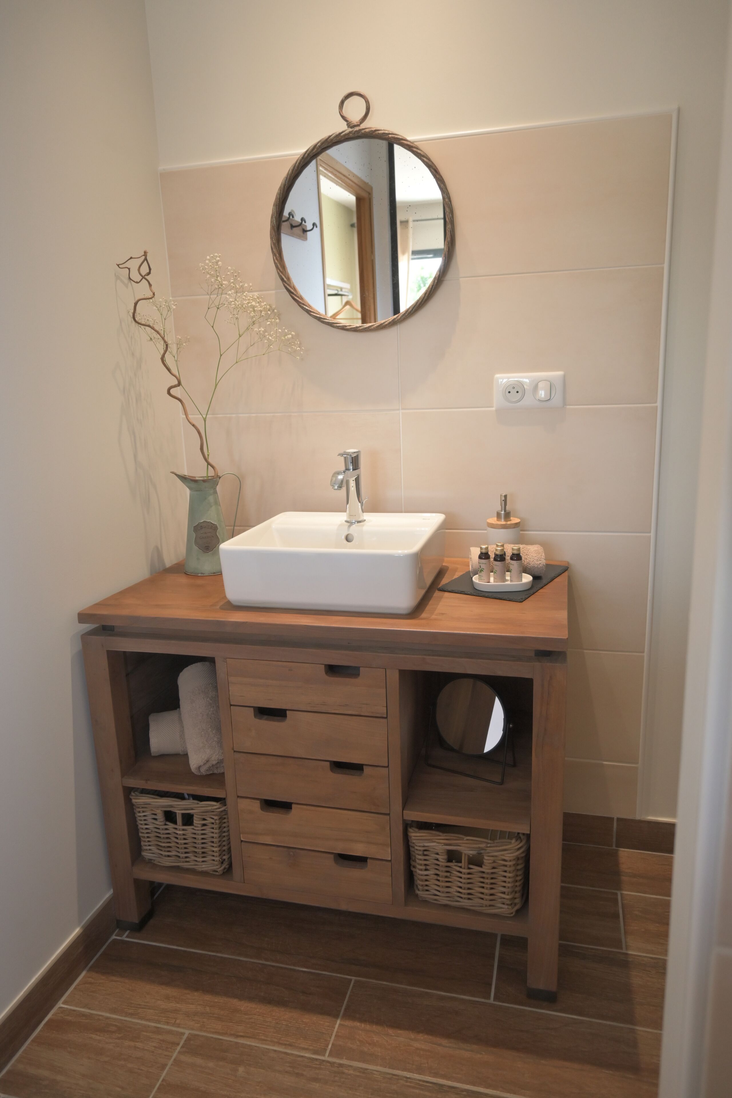 Une salle de bain avec un meuble-lavabo en bois, un lavabo blanc, un miroir et un vase avec des fleurs.
