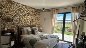 A bedroom with a stone wall, a bed with multiple pillows, a nightstand, a chair, a hanging light fixture, and a view of a green landscape through a sliding door. - Aux 3 Forets (Bizeneuille)