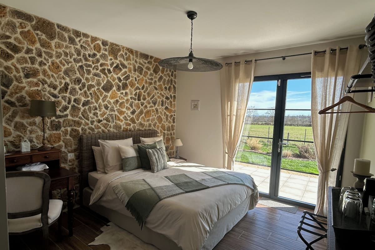 Une chambre avec un mur en pierre, un lit recouvert de plusieurs coussins, une table de chevet, une chaise, un luminaire suspendu et une vue sur un paysage verdoyant, visible à travers une porte coulissante.