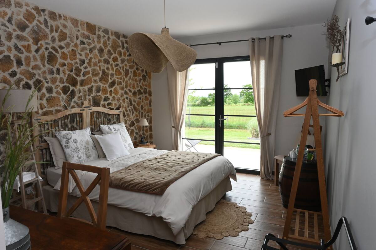 Une chambre avec un mur en pierre, un lit doté d’une tête de lit en bois, un luminaire suspendu et une vue sur l’extérieur grâce à une porte-fenêtre coulissante.